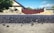 A close-up view of a freshly laid asphalt road, showcasing the rough texture and granular composition of the material. In the background, several structures are visible, including a beige house with a dark roof, a red fence, and construction mounds. The scene is set outdoors with clear weather and a blue sky featuring a few clouds.