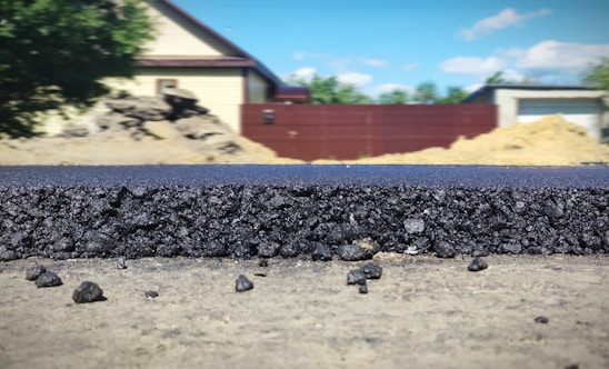 A close-up view of a freshly laid asphalt road, showcasing the rough texture and granular composition of the material. In the background, several structures are visible, including a beige house with a dark roof, a red fence, and construction mounds. The scene is set outdoors with clear weather and a blue sky featuring a few clouds.