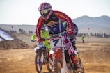 Several motocross riders are racing on a dirt track. The rider in the foreground is wearing a colorful helmet and riding a bike with the number 7. Dust is kicked up from the trail, and the background features a few spectators and mountainous terrain.