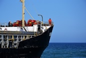 A large cargo ship with a black hull and white upper structures is visible with equipment and two crew members on deck. The ship is floating on a calm blue sea, and the sky is clear with no clouds.