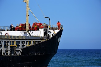 A large cargo ship with a black hull and white upper structures is visible with equipment and two crew members on deck. The ship is floating on a calm blue sea, and the sky is clear with no clouds.