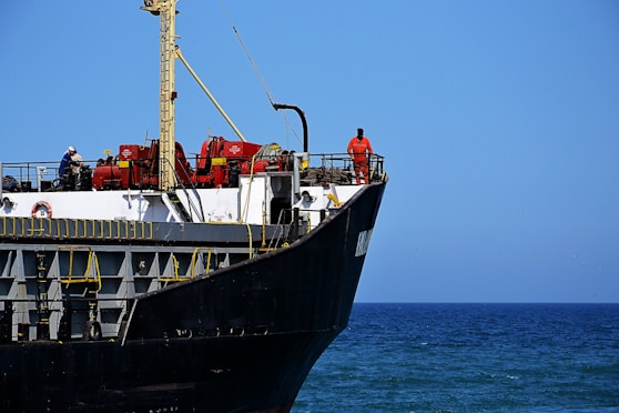 A large cargo ship with a black hull and white upper structures is visible with equipment and two crew members on deck. The ship is floating on a calm blue sea, and the sky is clear with no clouds.