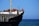 A large cargo ship with a black hull and white upper structures is visible with equipment and two crew members on deck. The ship is floating on a calm blue sea, and the sky is clear with no clouds.