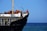 A large cargo ship with a black hull and white upper structures is visible with equipment and two crew members on deck. The ship is floating on a calm blue sea, and the sky is clear with no clouds.