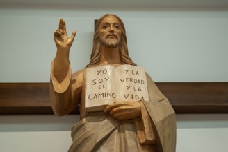 A wooden statue of a figure resembling Jesus Christ holding an open book with Spanish text. The inscription on the book reads 'YO SOY EL CAMINO Y LA VERDAD Y LA VIDA'. The statue features long hair and a bearded face, with an outstretched hand making a gesture. It stands against a wooden cross or background.