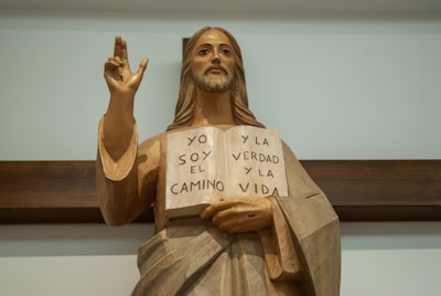 A wooden statue of a figure resembling Jesus Christ holding an open book with Spanish text. The inscription on the book reads 'YO SOY EL CAMINO Y LA VERDAD Y LA VIDA'. The statue features long hair and a bearded face, with an outstretched hand making a gesture. It stands against a wooden cross or background.