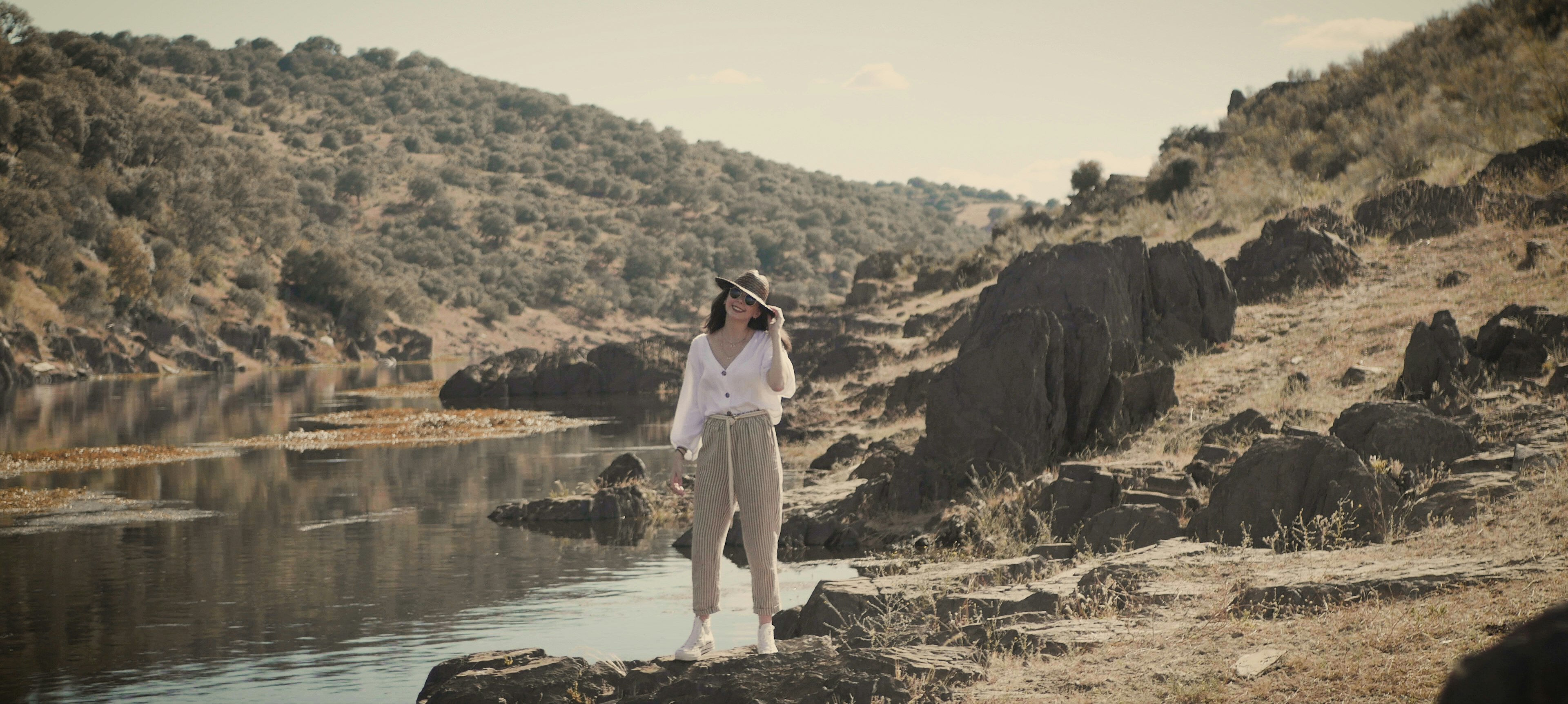 woman in white shirt and white pants standing on brown rock near river during daytime
