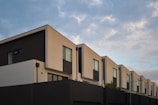 A row of modern, minimalist townhouses with flat roofs and geometric shapes. The buildings feature dark and light contrasting color panels with large rectangular windows. The sky is partly cloudy, providing a serene backdrop.