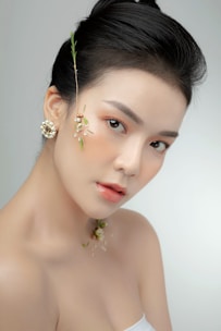 A woman with styled black hair is adorned with delicate floral accents on her face and neck. Her makeup is natural and soft, enhancing her features with a subtle elegance. A decorative earring complements the overall aesthetic. The background is a light, neutral tone, emphasizing the subject.