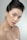 A woman with styled black hair is adorned with delicate floral accents on her face and neck. Her makeup is natural and soft, enhancing her features with a subtle elegance. A decorative earring complements the overall aesthetic. The background is a light, neutral tone, emphasizing the subject.