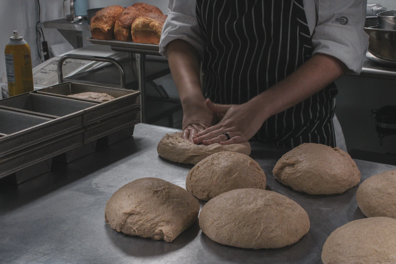 A baker in a striped apron shaping rounds of dough along a steel bench, finished loaves behind