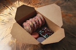 An open cardboard box contains a variety of colorful baked goods. Inside, there are cookies covered with rainbow sprinkles, a chocolate-covered brownie with colorful candy sprinkles, and two macarons, one pink and one purple. The box is set on a wooden table with a decorative pattern.