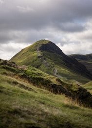 A lush green mountain peak bathed in sunlight, surrounded by a dramatic sky with clouds. A narrow trail winds up the ridge to the summit, where a small group of people can be seen silhouetted against the sky, indicating hikers reaching the top.