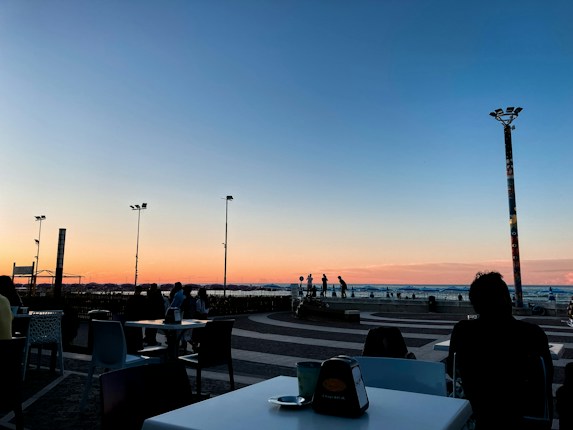 A serene coastal view during sunset with a gradient sky transitioning from warm orange near the horizon to a deep blue above. People are seated at outdoor tables of a caf&eacute;, and silhouettes of individuals are seen walking near the shore. Tall decorative lampposts are visible, adding to the scenic atmosphere.