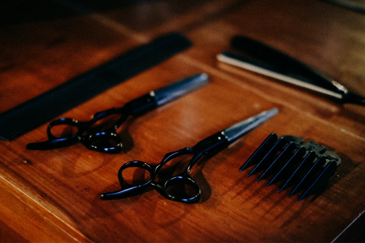 Two pairs of black hair scissors, a comb, a clipper guard and a straight razor laid out on a wooden table