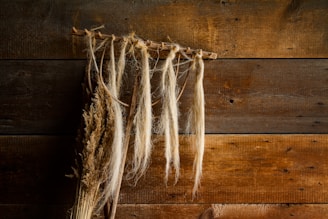 Dried plants and long strands of fibers hang from a rustic wooden beam against a backdrop of weathered wooden planks.