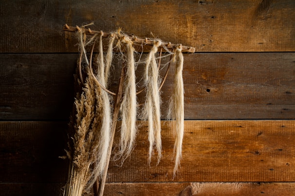 Dried plants and long strands of fibers hang from a rustic wooden beam against a backdrop of weathered wooden planks.