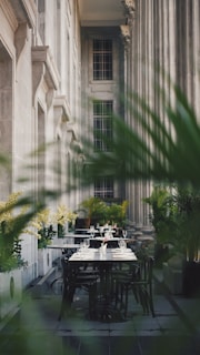 An elegant outdoor dining area is set amidst towering classical columns and lush greenery. The tables are neatly arranged with white tablecloths, sparkling glassware, and neatly folded napkins, creating an inviting atmosphere.
