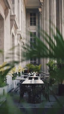 An elegant outdoor dining area is set amidst towering classical columns and lush greenery. The tables are neatly arranged with white tablecloths, sparkling glassware, and neatly folded napkins, creating an inviting atmosphere.