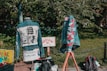 Two denim jackets are displayed outdoors, each decorated with messages and symbols related to social justice. The jacket on the left features a graphic with the text 'Black Lives Matter'. The jacket on the right displays various embroidered names and phrases. Signs nearby indicate that patches are available, and protest messages are visible. A green leafy background frames the scene.