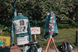 Two denim jackets are displayed outdoors, each decorated with messages and symbols related to social justice. The jacket on the left features a graphic with the text 'Black Lives Matter'. The jacket on the right displays various embroidered names and phrases. Signs nearby indicate that patches are available, and protest messages are visible. A green leafy background frames the scene.