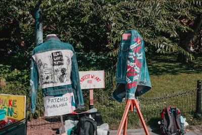 Two denim jackets are displayed outdoors, each decorated with messages and symbols related to social justice. The jacket on the left features a graphic with the text 'Black Lives Matter'. The jacket on the right displays various embroidered names and phrases. Signs nearby indicate that patches are available, and protest messages are visible. A green leafy background frames the scene.