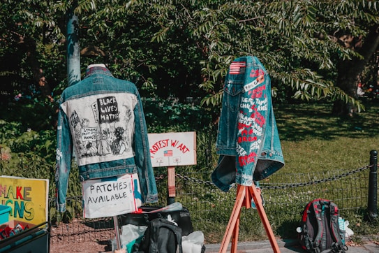 Two denim jackets are displayed outdoors, each decorated with messages and symbols related to social justice. The jacket on the left features a graphic with the text 'Black Lives Matter'. The jacket on the right displays various embroidered names and phrases. Signs nearby indicate that patches are available, and protest messages are visible. A green leafy background frames the scene.