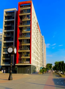 A modern, multi-story apartment building with red accents on the balconies is situated along a wide, paved pedestrian pathway. There is also a decorative street clock with two faces showing different time zones. On the right, some people are walking along the path lined with trees and parked cars.