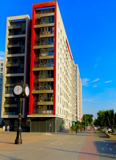 A modern, multi-story apartment building with red accents on the balconies is situated along a wide, paved pedestrian pathway. There is also a decorative street clock with two faces showing different time zones. On the right, some people are walking along the path lined with trees and parked cars.
