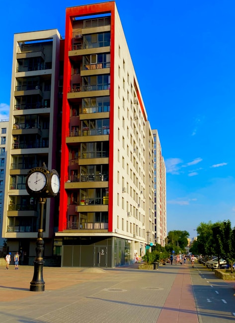 A modern, multi-story apartment building with red accents on the balconies is situated along a wide, paved pedestrian pathway. There is also a decorative street clock with two faces showing different time zones. On the right, some people are walking along the path lined with trees and parked cars.
