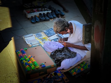 An elderly person is seated on the ground, surrounded by various goods laid out for sale, such as colorful toy frogs, small packages, and shoes. The individual is wearing a white shirt and is focused on working with their hands. There is a mixture of sunlight and shadow on the pavement where the items are displayed.