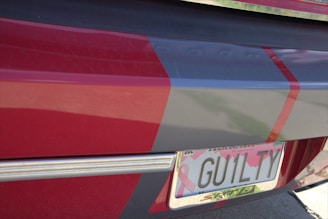 A close-up of a car's rear featuring a personalized license plate with the text 'GUILTY'. The vehicle is painted with red and gray colors, and the plate has a pink ribbon design, commonly associated with breast cancer awareness.