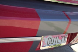 A close-up of a car's rear featuring a personalized license plate with the text 'GUILTY'. The vehicle is painted with red and gray colors, and the plate has a pink ribbon design, commonly associated with breast cancer awareness.