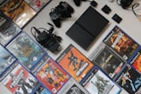 A collection of PlayStation 2 video games and accessories, including a console, several game controllers, power cables, and a variety of game cases. The game titles include popular names like 'Lord of the Rings' and 'Metal Gear Solid 2'. The items are neatly arranged on a light-colored surface with the game cases forming a rectangle around the console and accessories.