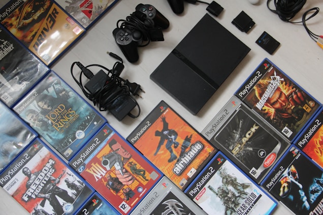 A collection of PlayStation 2 video games and accessories, including a console, several game controllers, power cables, and a variety of game cases. The game titles include popular names like 'Lord of the Rings' and 'Metal Gear Solid 2'. The items are neatly arranged on a light-colored surface with the game cases forming a rectangle around the console and accessories.