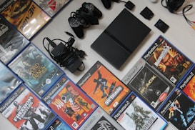 A collection of PlayStation 2 video games and accessories, including a console, several game controllers, power cables, and a variety of game cases. The game titles include popular names like 'Lord of the Rings' and 'Metal Gear Solid 2'. The items are neatly arranged on a light-colored surface with the game cases forming a rectangle around the console and accessories.