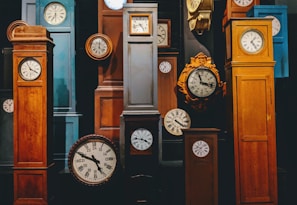A collection of variously styled grandfather and wall clocks are arranged closely together. The clocks display traditional Roman numerals and classic clock faces. The wooden frames come in different shades of brown, with some metal parts and intricate designs.