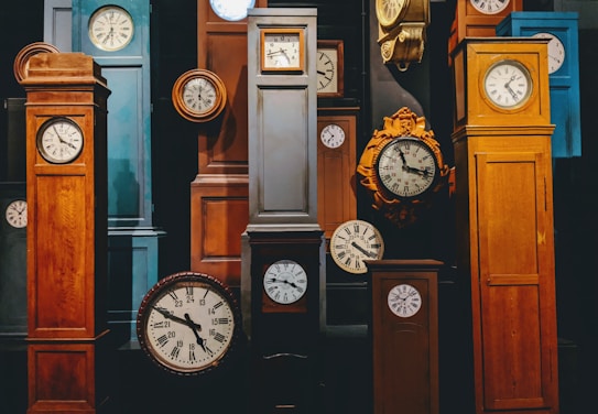 A collection of variously styled grandfather and wall clocks are arranged closely together. The clocks display traditional Roman numerals and classic clock faces. The wooden frames come in different shades of brown, with some metal parts and intricate designs.