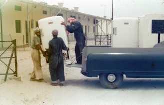 A group of men are unloading large white containers from the back of a blue pickup truck. The scene appears to be set in a dusty outdoor area adjacent to a building with numerous windows and a metal fence. The men are wearing work clothes and head coverings, and they are working together to maneuver the containers.
