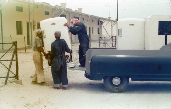 A group of men are unloading large white containers from the back of a blue pickup truck. The scene appears to be set in a dusty outdoor area adjacent to a building with numerous windows and a metal fence. The men are wearing work clothes and head coverings, and they are working together to maneuver the containers.