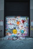 A garage door covered in colorful graffiti including various symbols, words like 'REWARD' and 'CULTURAL', smiley faces, hearts, and abstract shapes. Two no parking signs are visible on either side.