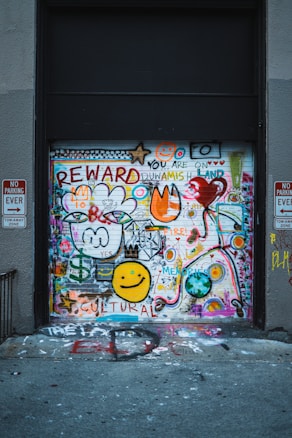 A garage door covered in colorful graffiti including various symbols, words like 'REWARD' and 'CULTURAL', smiley faces, hearts, and abstract shapes. Two no parking signs are visible on either side.