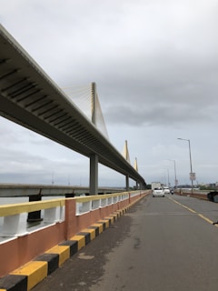 A broad view of a modern bridge with suspension cables and a roadway underneath. The sky is overcast, suggesting a cloudy day. Vehicles, including a white car and a motorcycle, are visible on the road. The bridge structure is primarily grey with yellow accents, and the road is bordered by safety barricades in yellow, black, and orange.
