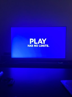 A television screen displays the phrase 'PLAY HAS NO LIMITS.' in bold white text against a blue background. The room is dimly lit with the screen being the primary light source, casting a blue hue on the surroundings. A table in front of the TV has indistinguishable objects that are partially visible in the foreground.