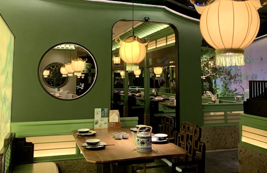 An elegantly designed restaurant interior featuring soft green walls and warm lighting from large hanging lamps. A wooden dining table is set with plates, bowls, and a teapot, surrounded by intricately designed chairs. A circular mirror on the wall reflects the ambient light, creating a cozy and inviting atmosphere. Decorative elements, including plants and artistic wall designs, enhance the calming environment.