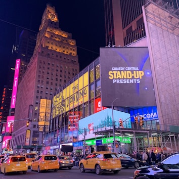 A bustling urban scene at night with towering buildings illuminated by vibrant neon lights and digital displays. Large advertisements for 'The Lion King' musical and 'Comedy Central Stand-Up Presents' are prominently featured. Several yellow taxis and vehicles are moving through a busy intersection, and people can be seen on the sidewalks.