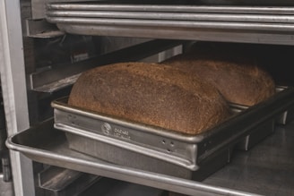 Two loaves of bread are placed in a metal baking pan on an oven rack. The bread has a dark, golden-brown crust, and the pans are labeled 'Chicago Metallic'. The setting appears to be an industrial kitchen or bakery.