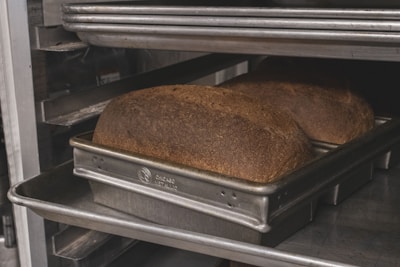 Two loaves of bread are placed in a metal baking pan on an oven rack. The bread has a dark, golden-brown crust, and the pans are labeled 'Chicago Metallic'. The setting appears to be an industrial kitchen or bakery.