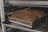 Two loaves of bread are placed in a metal baking pan on an oven rack. The bread has a dark, golden-brown crust, and the pans are labeled 'Chicago Metallic'. The setting appears to be an industrial kitchen or bakery.