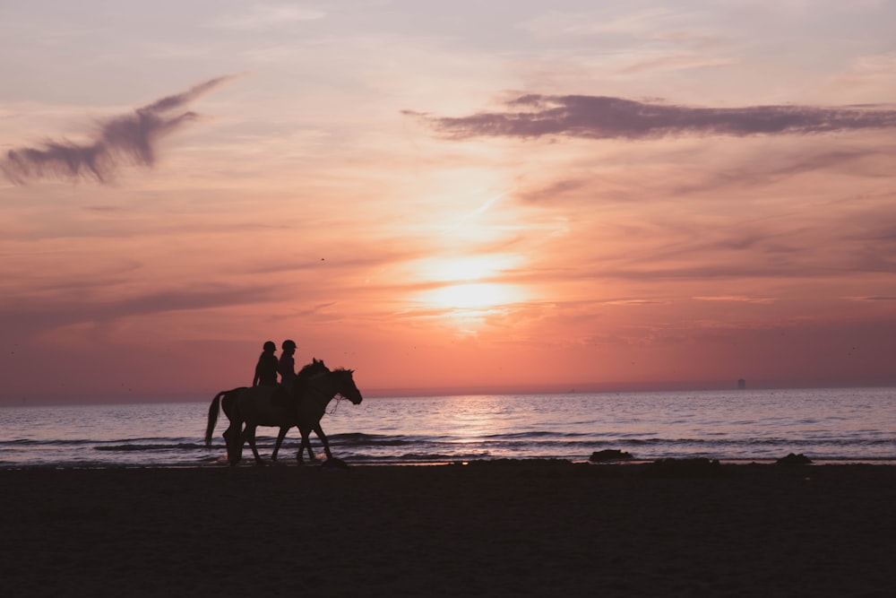 Silueta de 2 personas montando a caballo en la playa durante la puesta del sol